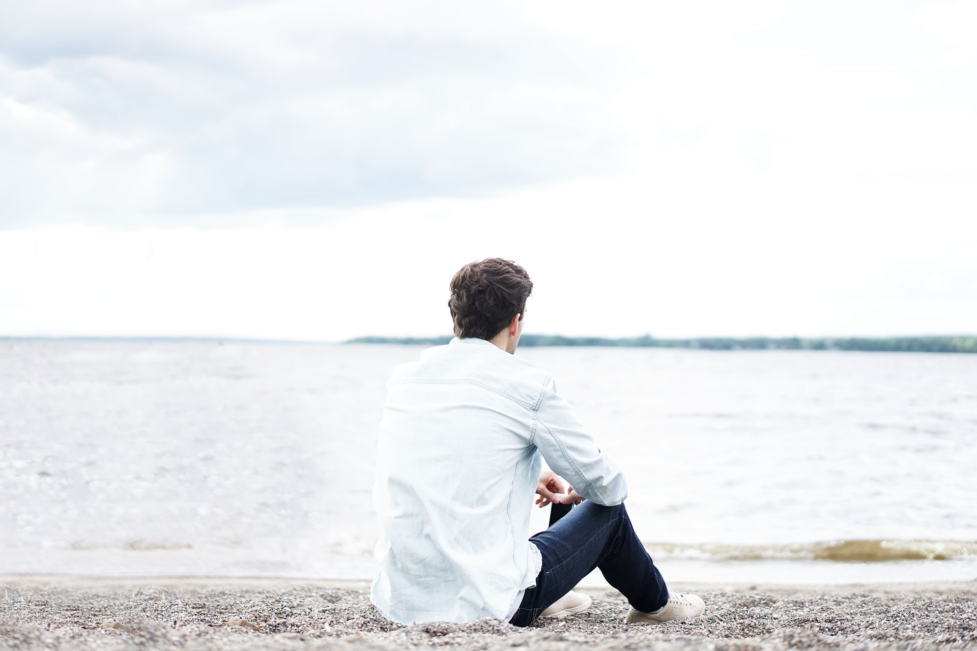 Mann in weißem Hemd sitzt am Strand und schaut auf das Meer