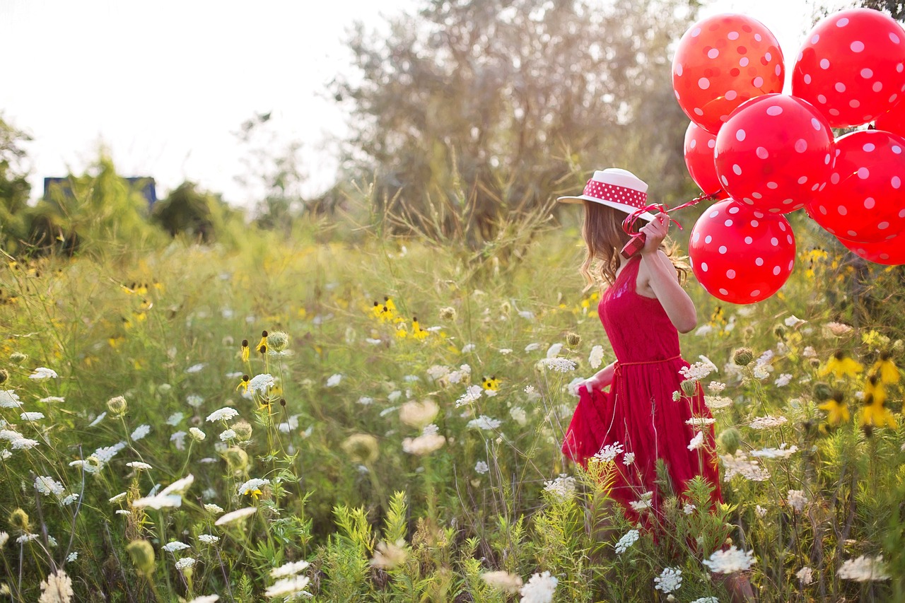 Frau mit Sonnenhut, rotem Kleid und rot getupften Luftballons in der Hand läuft durch eine Blumenwiese