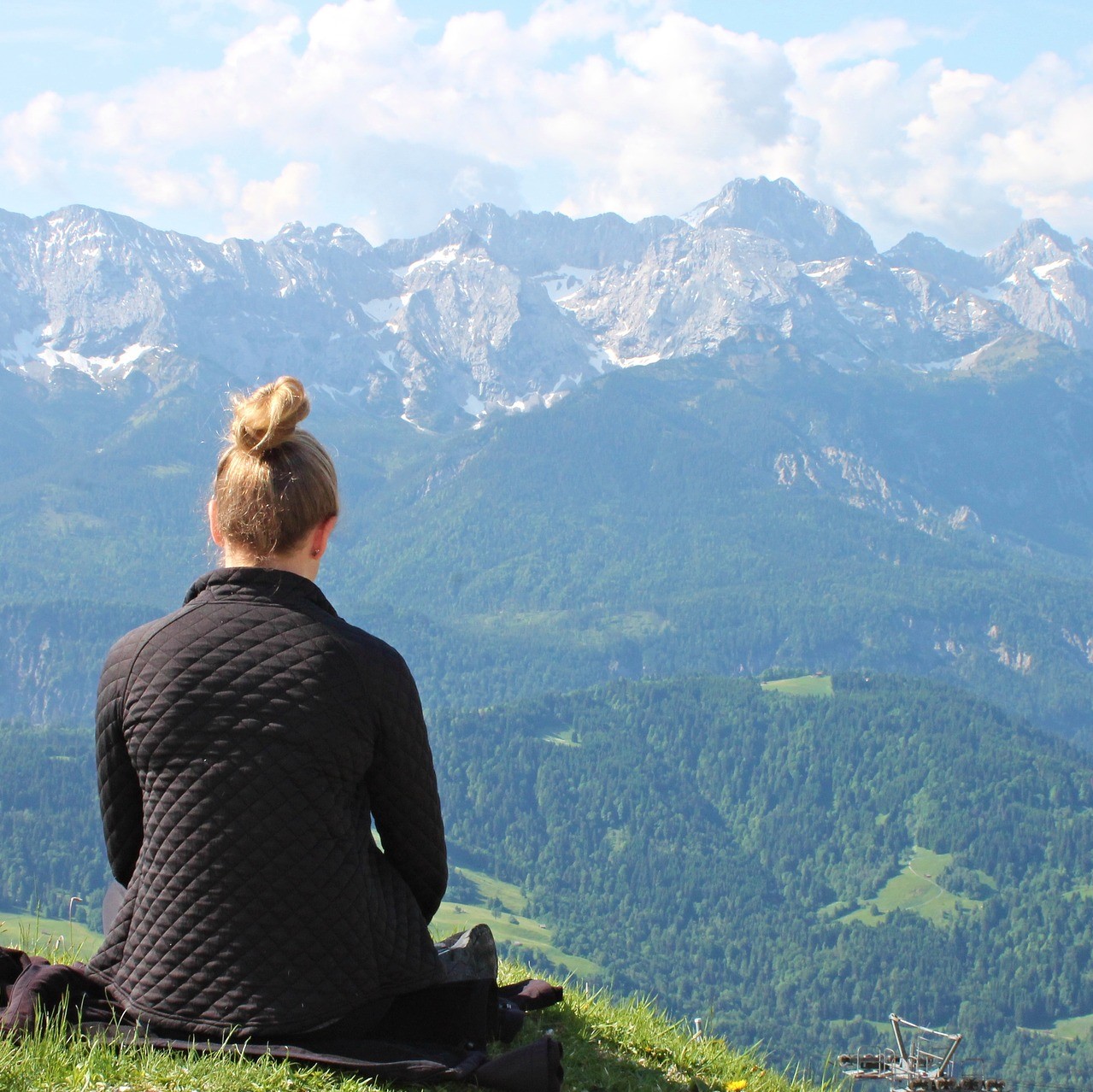 Frau sitzt auf einer Wiese und schaut auf die Berge und das Tal vor sich und erlebt es wie eine Meditation