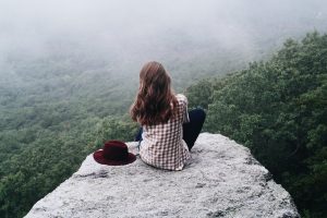 Frau von hinten zu sehen auf einem Felsblock, den Hut neben sich, sie schaut über Bäume und weiß, dass mit Mut die schönsten Geschichten anfangen